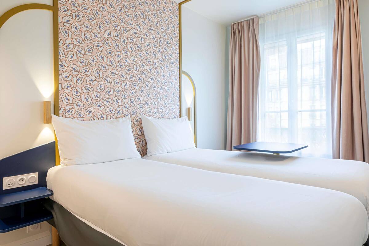 A double bed with a headboard, bedside tables, a power outlet, and a window with curtains.