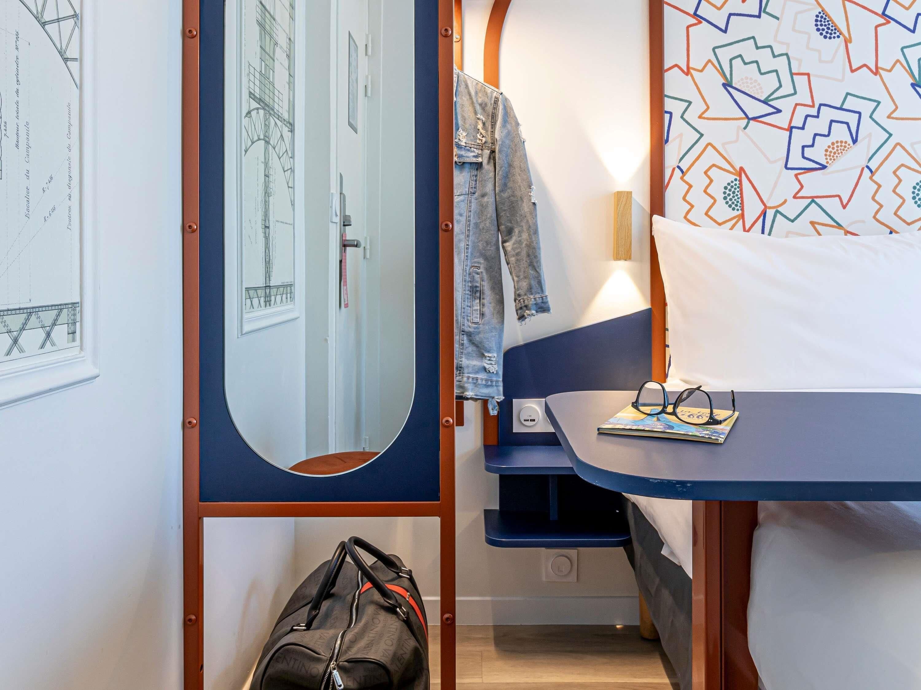 Um quarto com uma cama, uma mesa de cabeceira com copos, um casaco pendurado na porta e uma mala no chão.