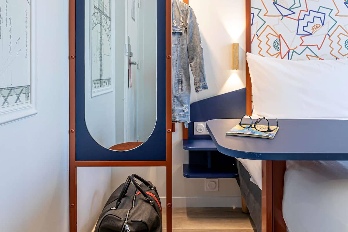 Um quarto com uma cama, uma mesa de cabeceira com copos, um casaco pendurado na porta e uma mala no chão.