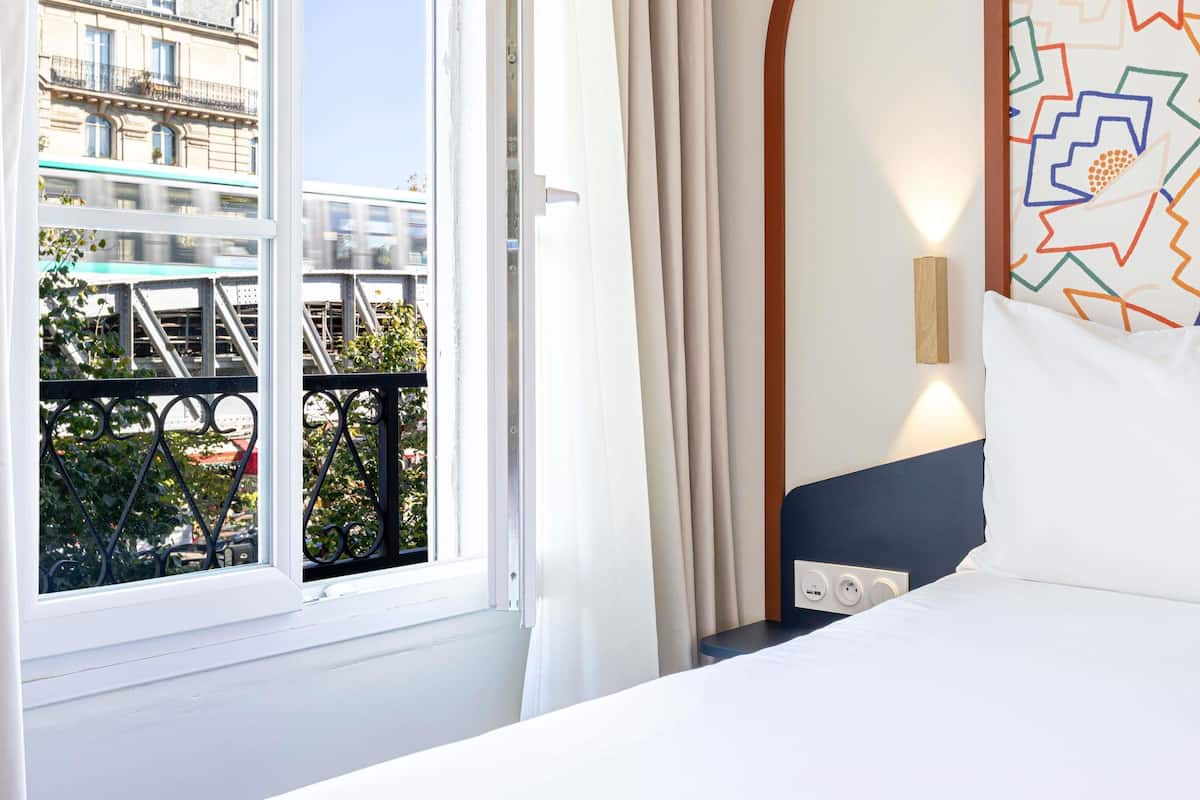 Une chambre d’hôtel avec une grande fenêtre, un lit, une table de chevet et un luminaire fixé au mur.