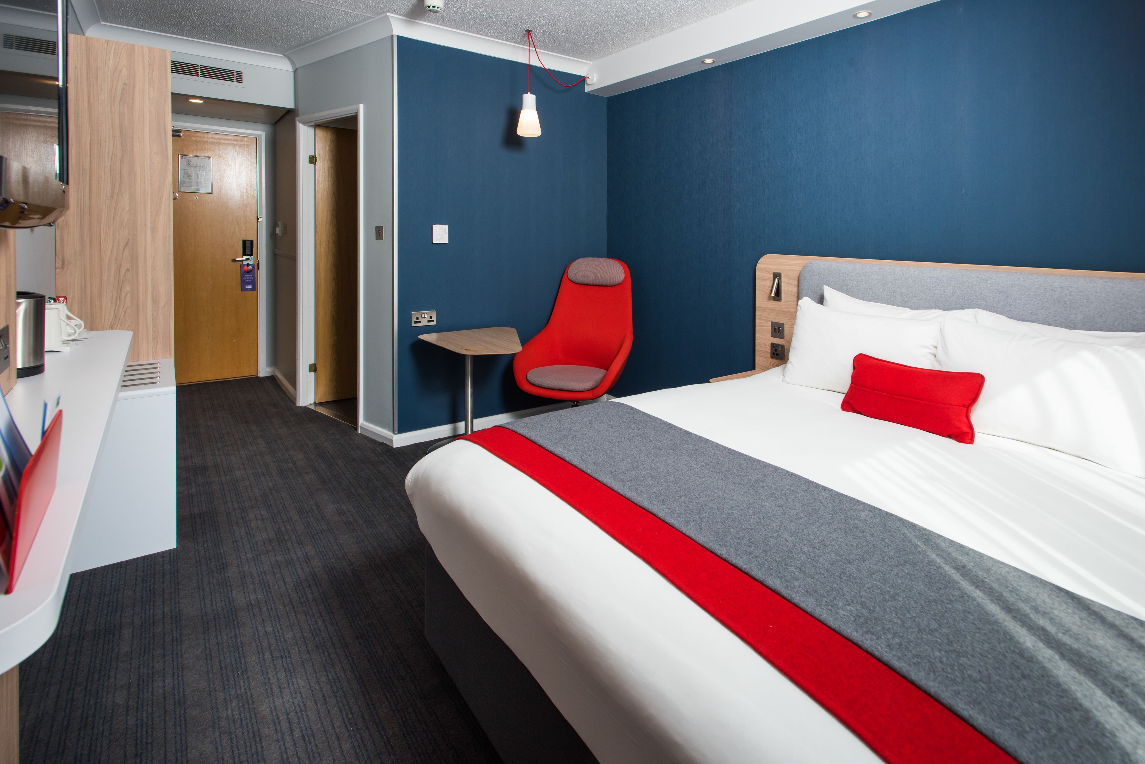 A hotel room with a large bed, a red chair, a small table, and a wooden door.