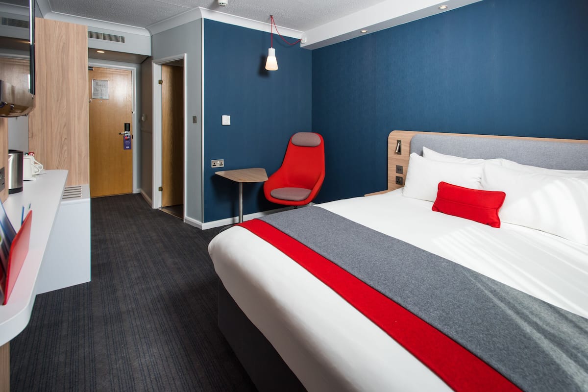 A hotel room with a large bed, a red chair, a small table, and a wooden door.