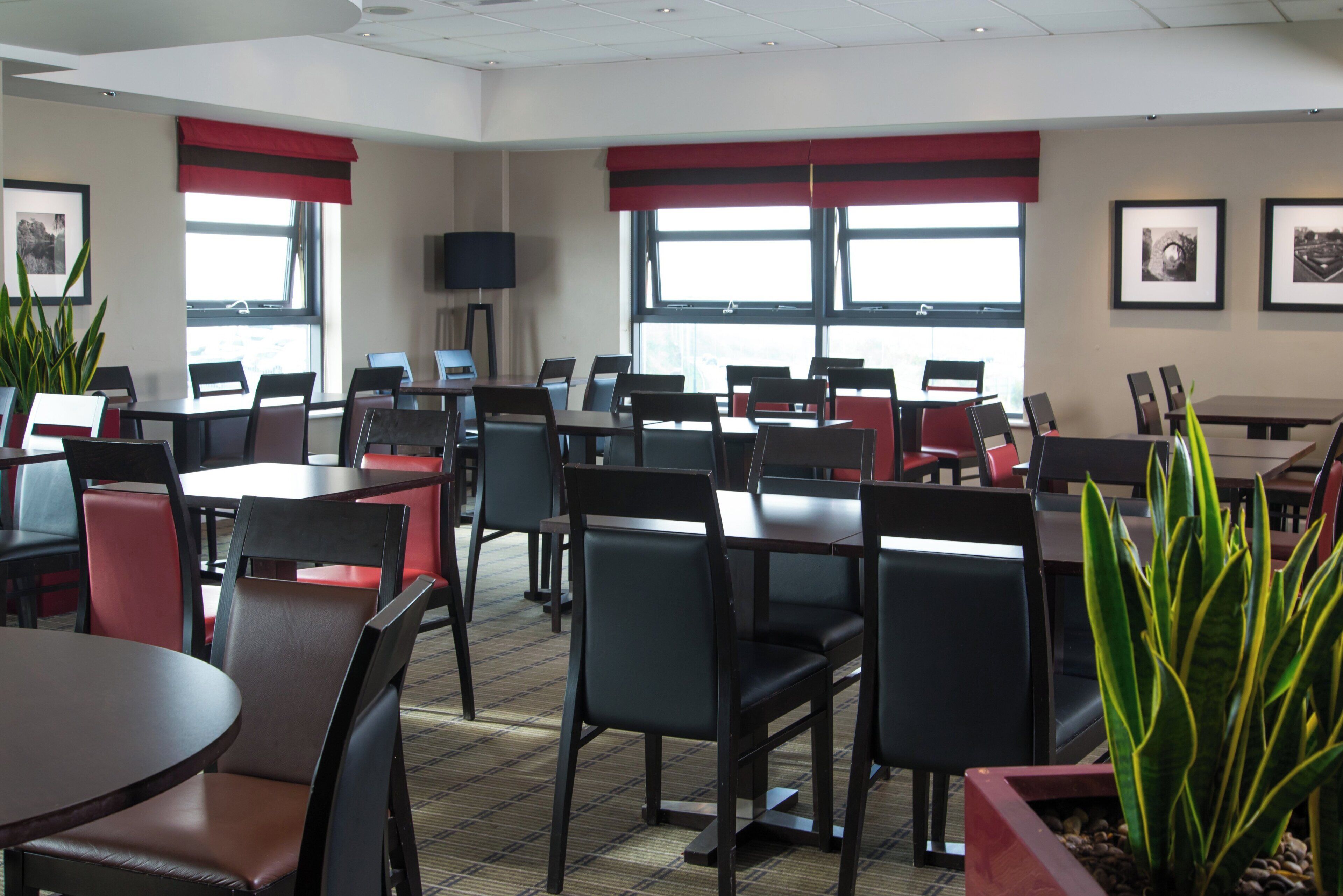 A hotel dining area with multiple tables and chairs, large windows with red curtains, and framed pictures on the walls.