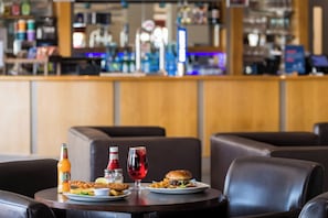 Una mesa de comedor con comida que incluye pescado con papas fritas, una hamburguesa y una copa de vino tinto, y un bar al fondo.