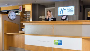 A Holiday Inn Express reception desk with a clock, a digital display, and a sign indicating the under 25 age group. - Holiday Inn Express London Luton Airport by IHG (Luton)