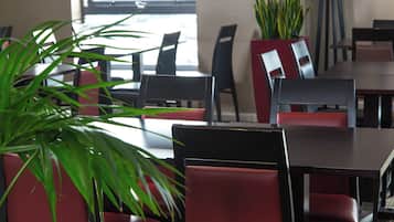 Interior de un restaurante con sillas rojas y negras, un ventanal grande y plantas en macetas.