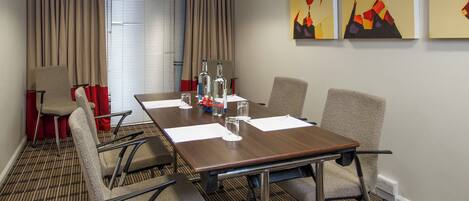A conference room with a long table, four chairs, and two windows with blinds.