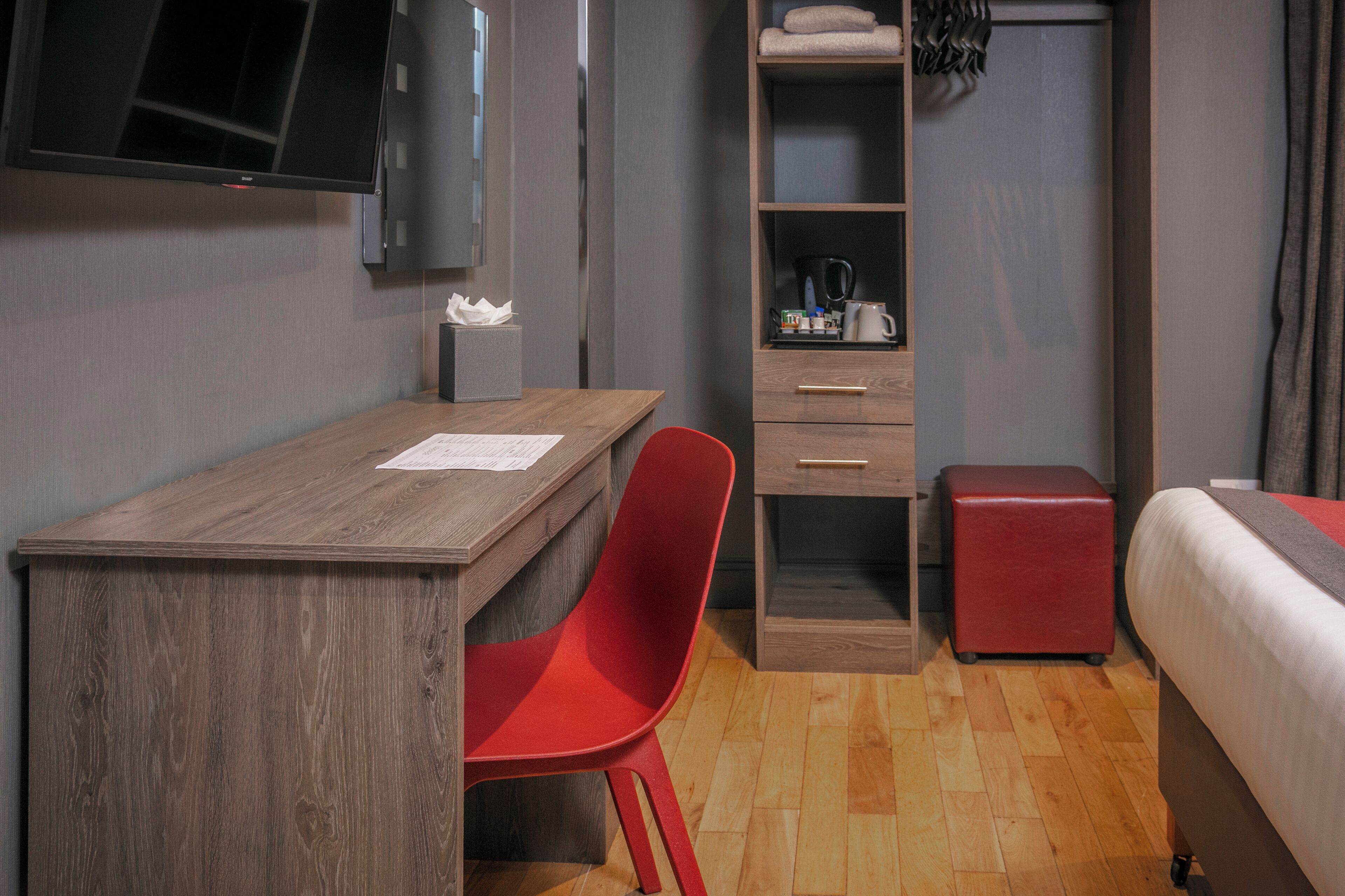 A hotel room with a wooden desk, a red chair, a bed, a wardrobe, and a television mounted on the wall.
