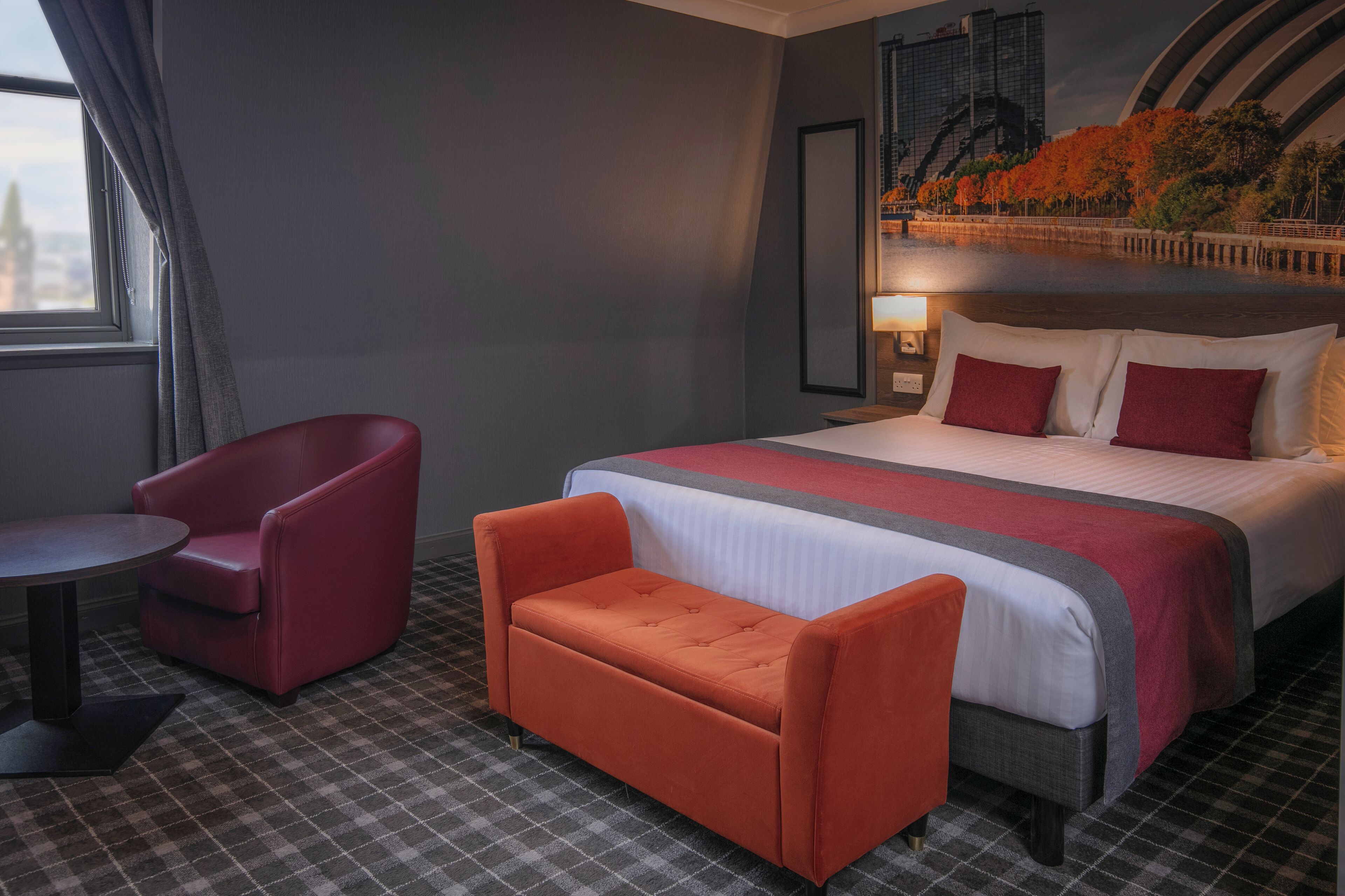 Une chambre d’hôtel avec un grand lit, un fauteuil rouge, un plus petit fauteuil orange et un sol à carreaux.