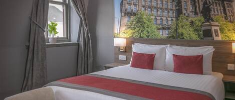 Habitación de hotel con una cama grande, ventana con cortinas, mesita de noche, lámpara y vista a un edificio con una estatua.