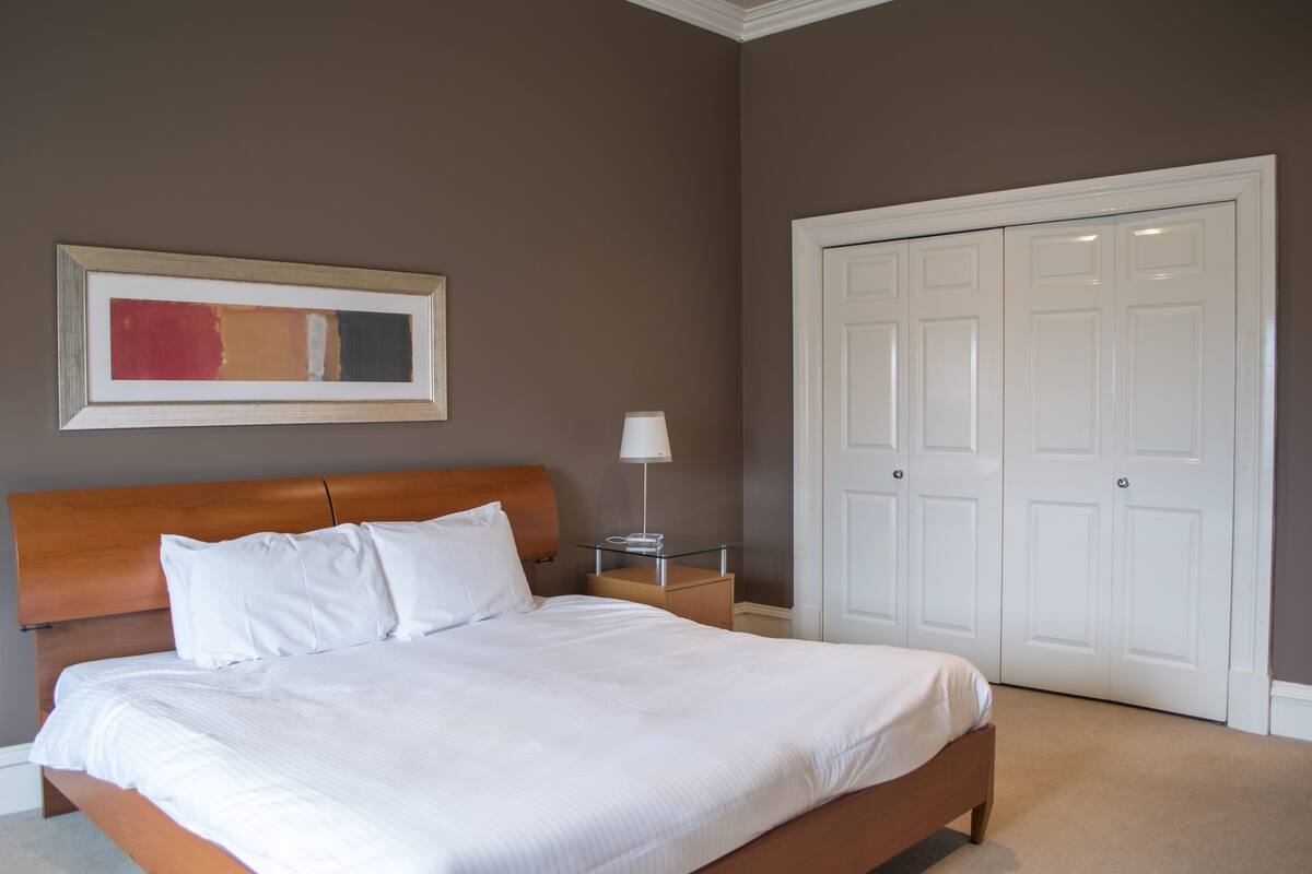 A neatly made bed with white linens, a wooden headboard, a bedside table with a lamp, a framed abstract painting, and a set of white double doors.