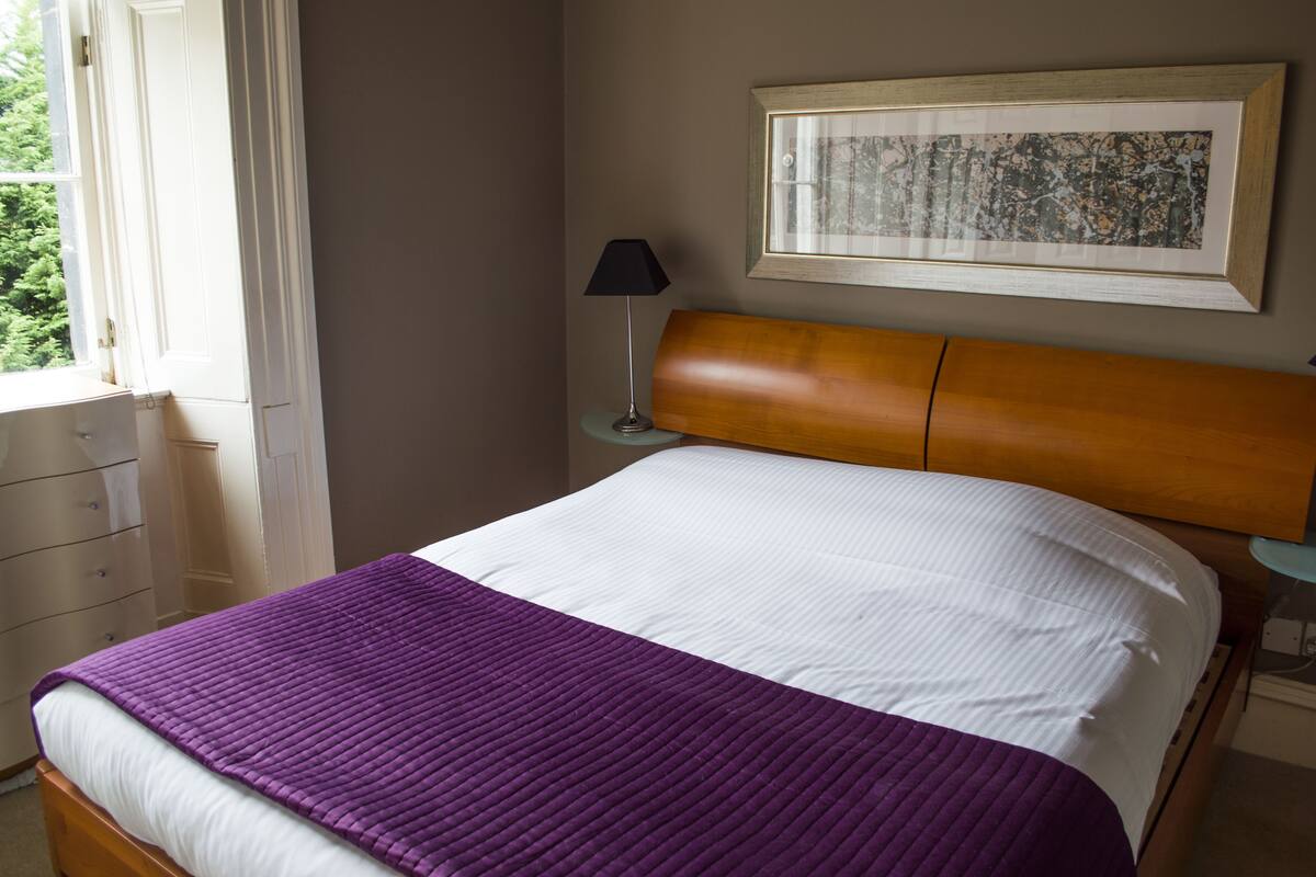 A neatly made bed with a white and purple bedspread, a wooden headboard, a framed picture on the wall, and a window with curtains.