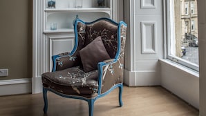 A room with a wooden floor, a white fireplace with shelves, a blue armchair, and a large window with a view of buildings. - Dreamhouse Apartments Glasgow West End (Glasgow)