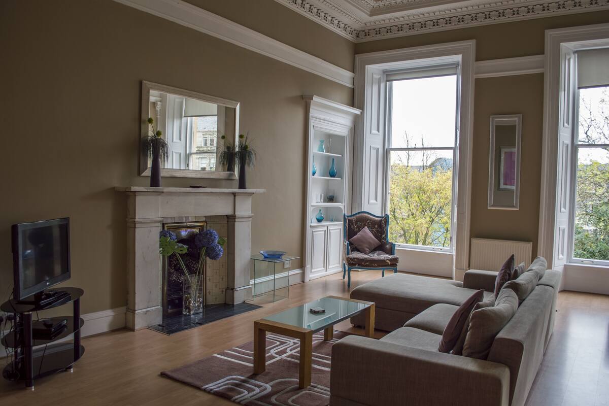 A living room with a fireplace, a television, a sofa, and a large window offering a view of trees.