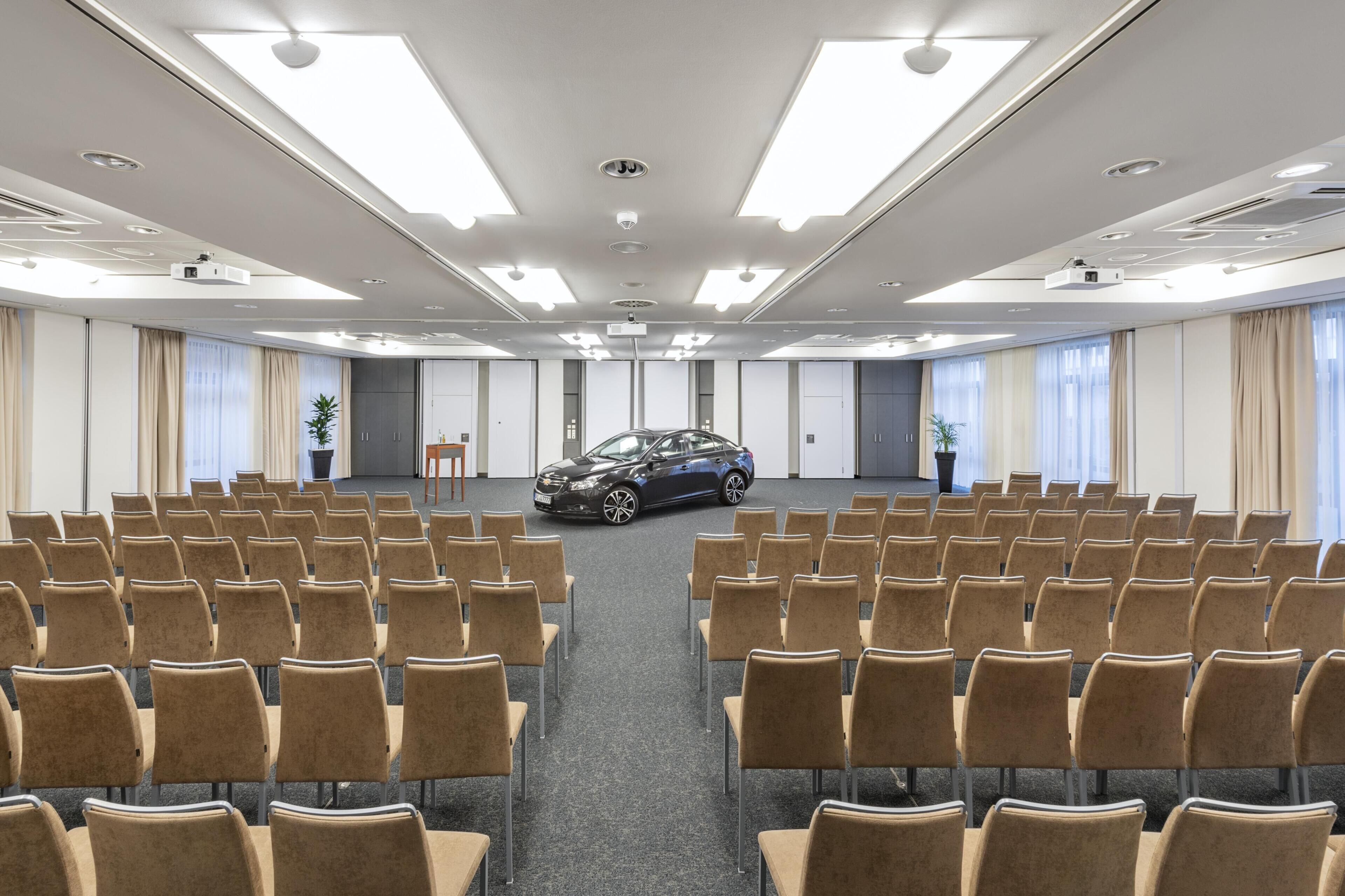 Un salón de conferencias con filas de sillas, un proyector y un automóvil estacionado en el centro.