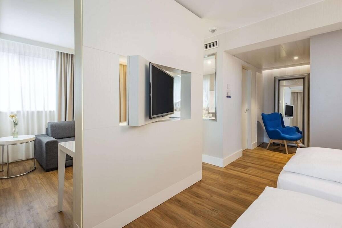 A hotel room with a flat-screen TV mounted on the wall, a grey sofa, a small round table with a vase, and a blue chair.