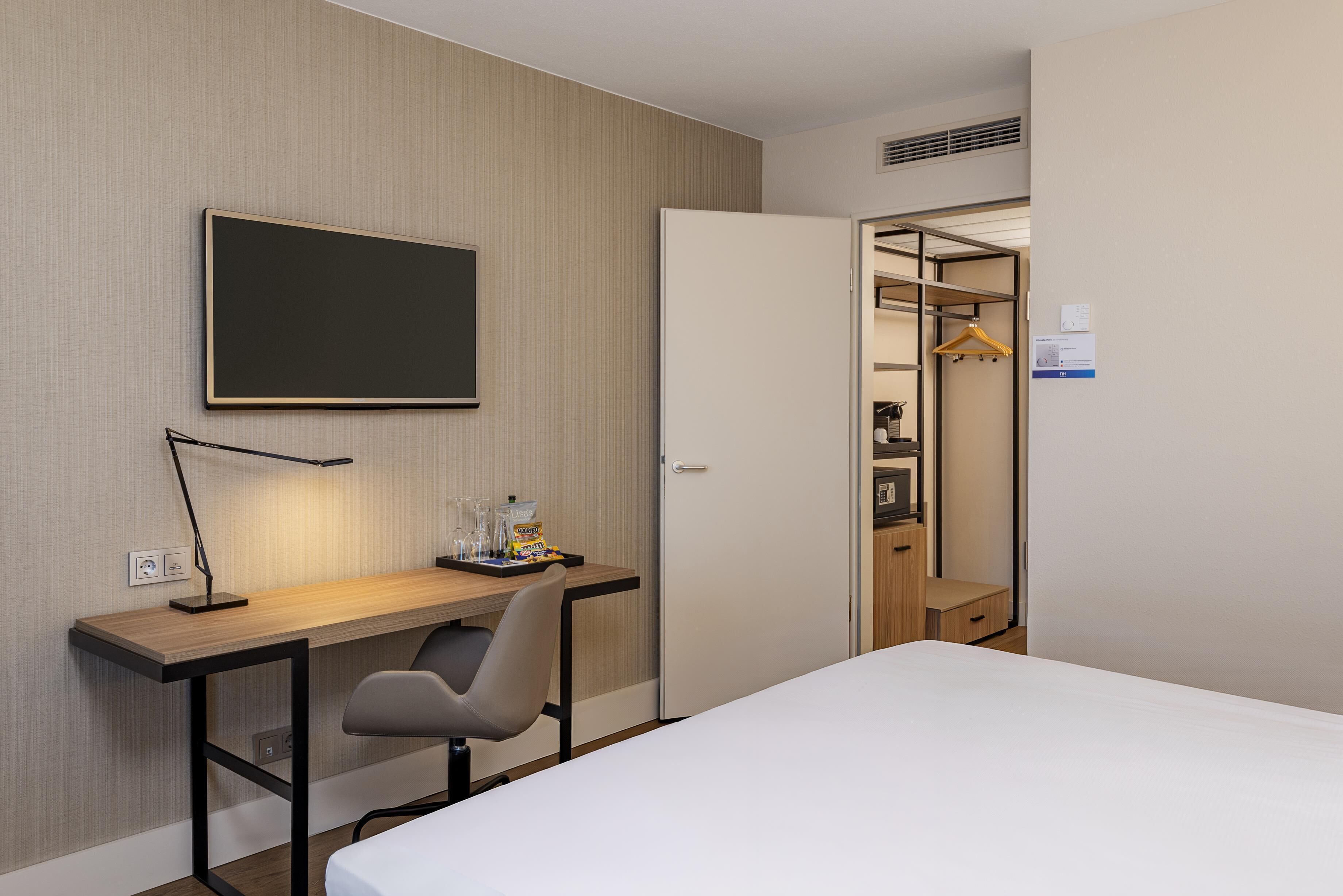 A hotel room with a bed, a desk with a lamp, a chair, a wardrobe, and a coat rack.