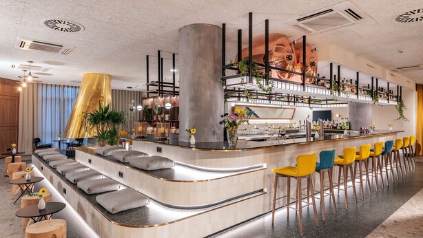 Un moderno bar con una barra curva, diversas opciones de asiento, incluidas sillas de bar y asientos acolchados, y una variedad de plantas y elementos decorativos.