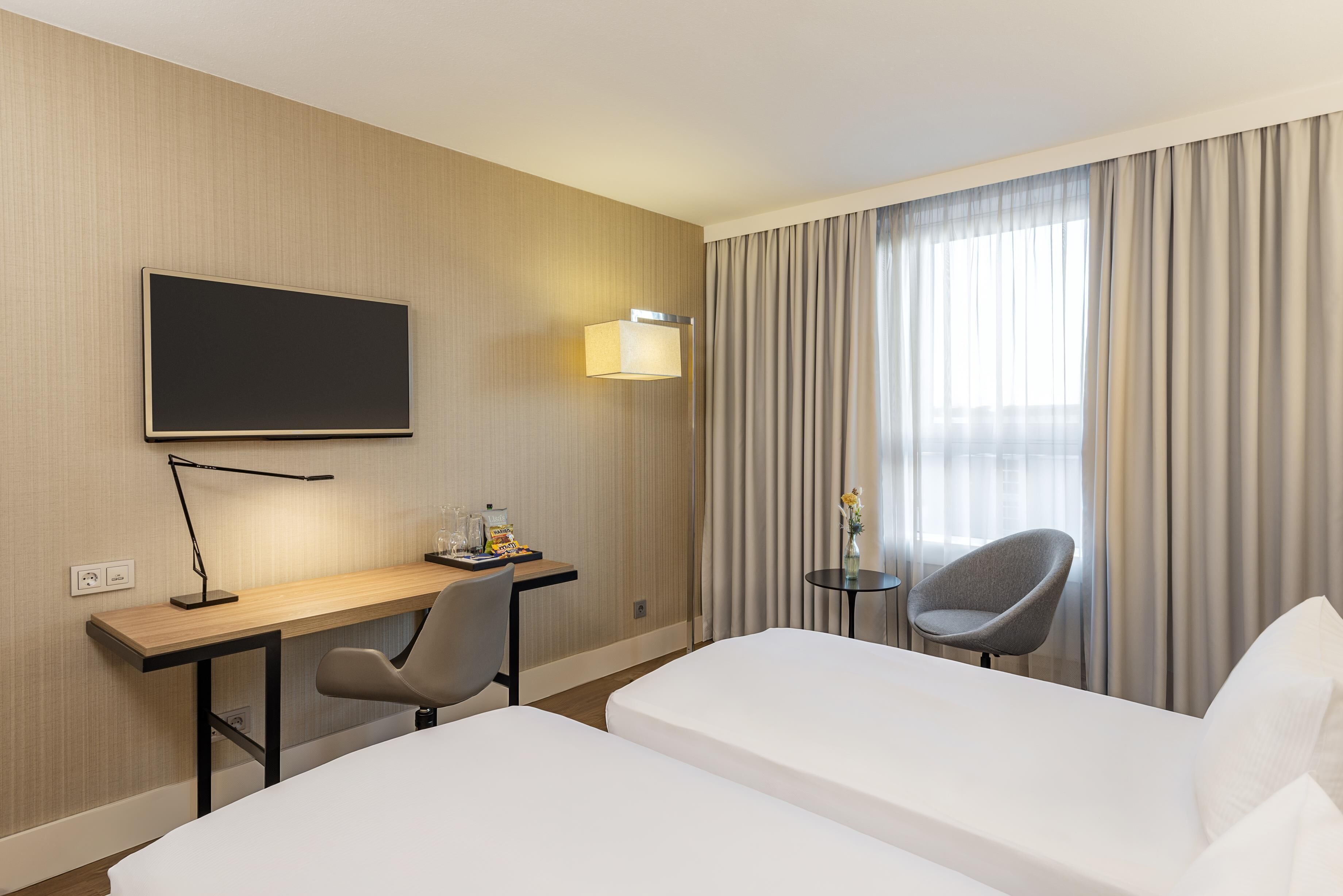 Ett hotellrum med ett skrivbord, två enkelsängar, en stol, en TV och ett fönster med gardiner.
