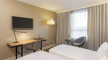 A hotel room with a desk, two single beds, a chair, a TV, and a window with curtains.