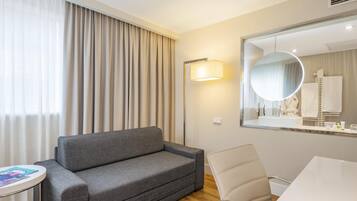 Een hotelkamer met een grijze bank, een witte stoel, een ronde spiegel en een klein tafeltje met een tijdschrift.