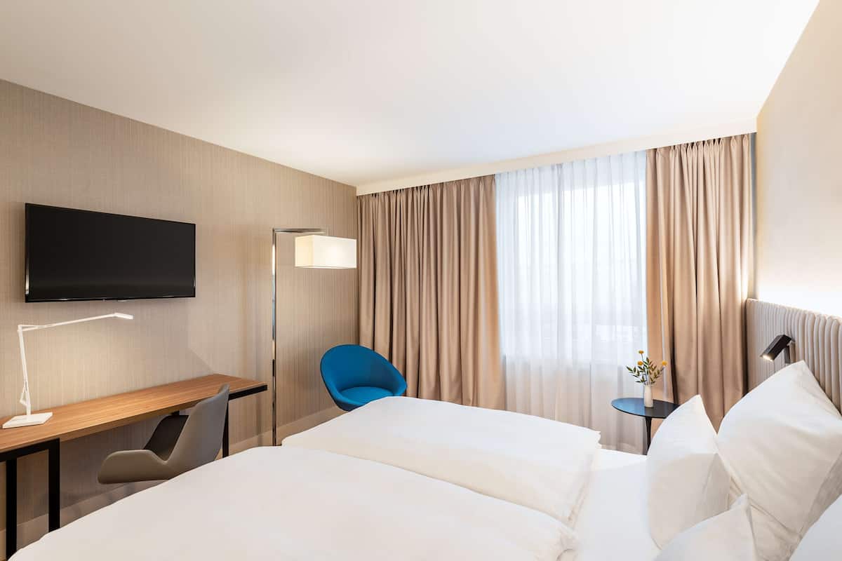 A hotel room with a large bed, a desk with a chair, a television mounted on the wall, and a small table with a lamp.