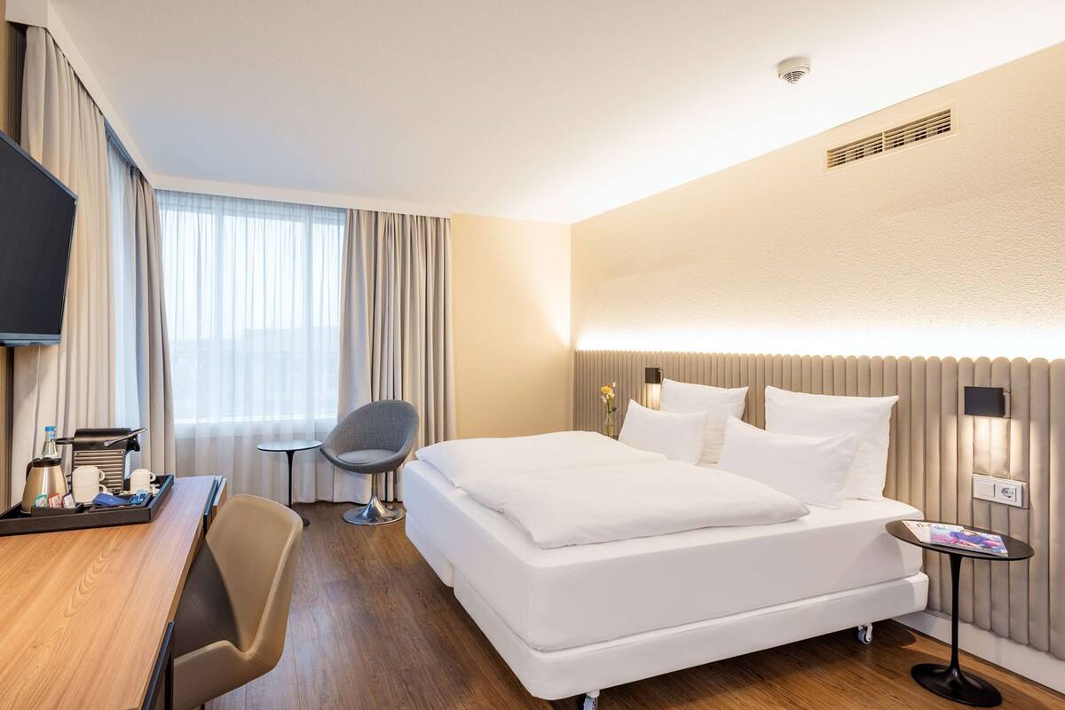 A hotel room with a bed, a desk with a chair, a TV, and a bedside table with a lamp.