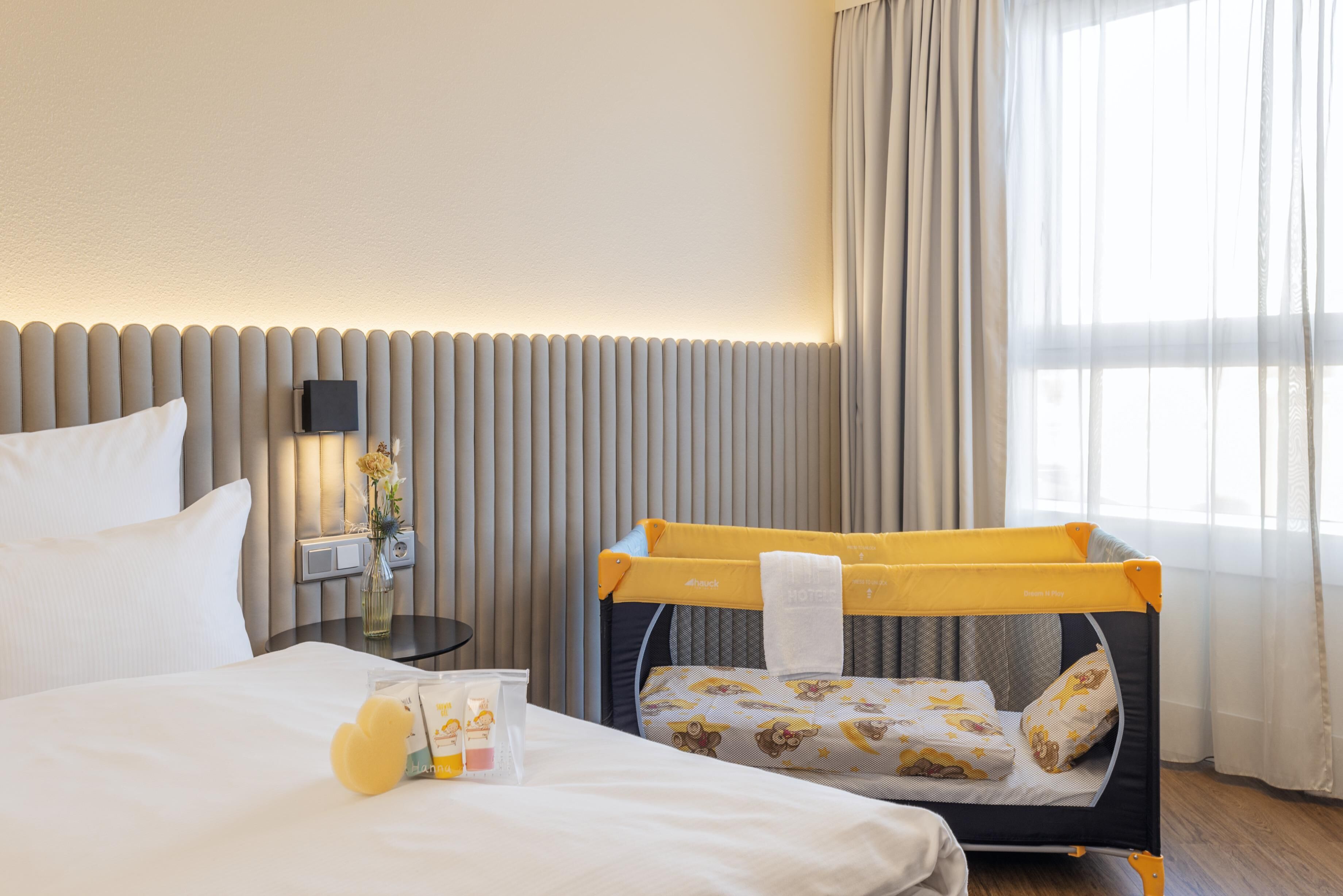 Ein Hotelzimmer mit Bett, Nachttisch, Lampe, einem Fenster mit Vorhängen und einem Reisebett.