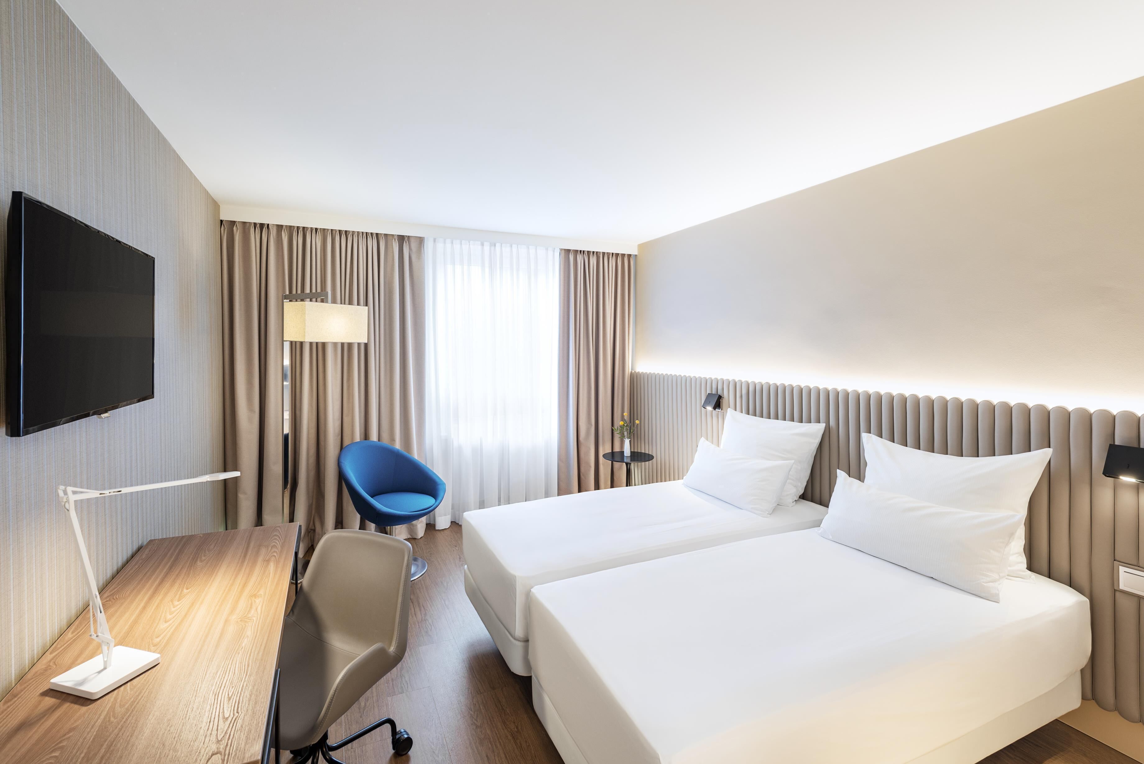 A hotel room with two beds, a desk with a chair, a TV, and a window with curtains.