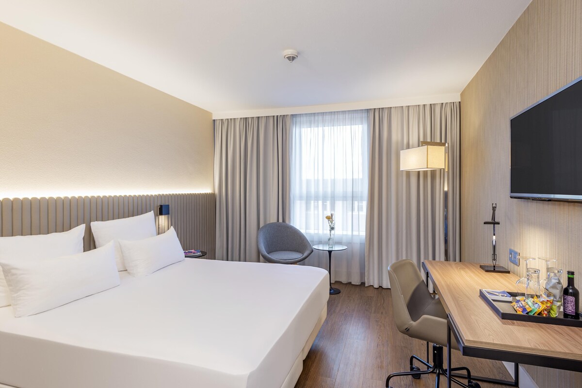 A hotel room with a large bed, a desk with a chair, a television, and a window with curtains.