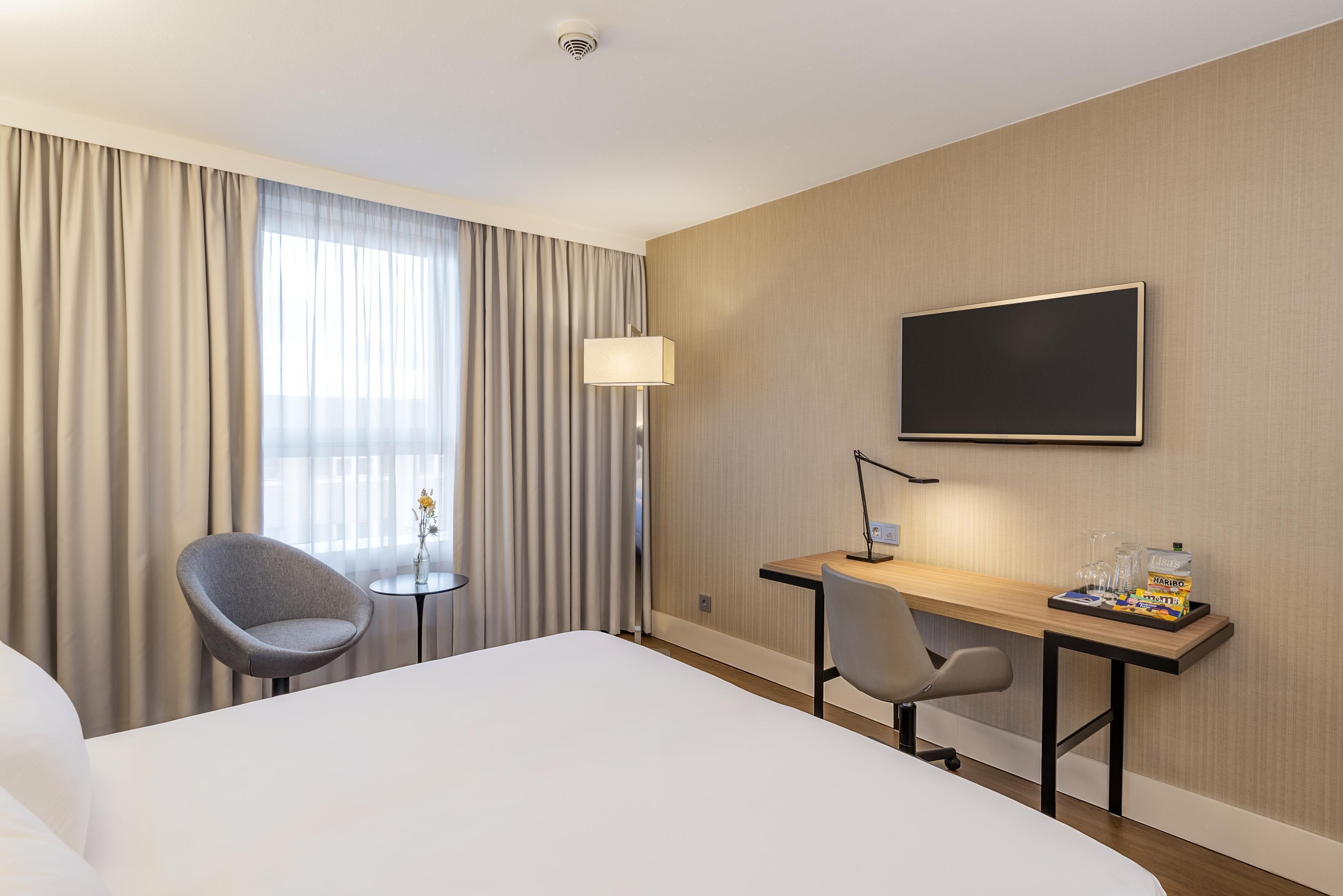 A hotel room with a bed, two chairs, a desk, a flat-screen TV, a lamp, and a window with curtains.