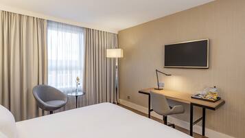 Habitación de hotel con cama, dos sillas, escritorio, televisor de pantalla plana, lámpara y ventana con cortinas.