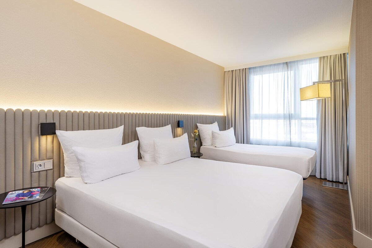 A hotel room with two beds, a bedside table, a lamp, and a window with curtains.