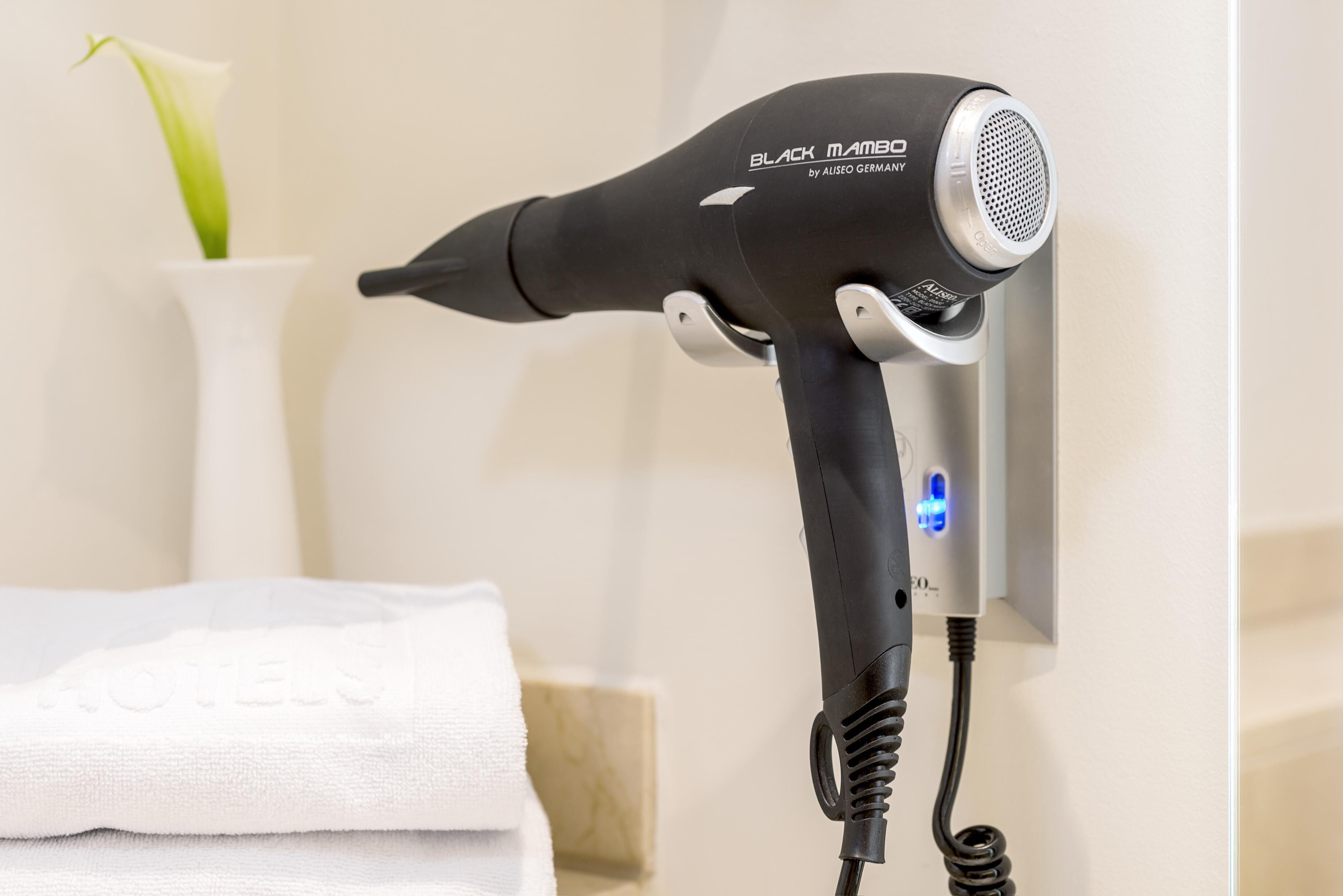 A hair dryer mounted on a wall, with a white towel and a vase with a green plant in the background.