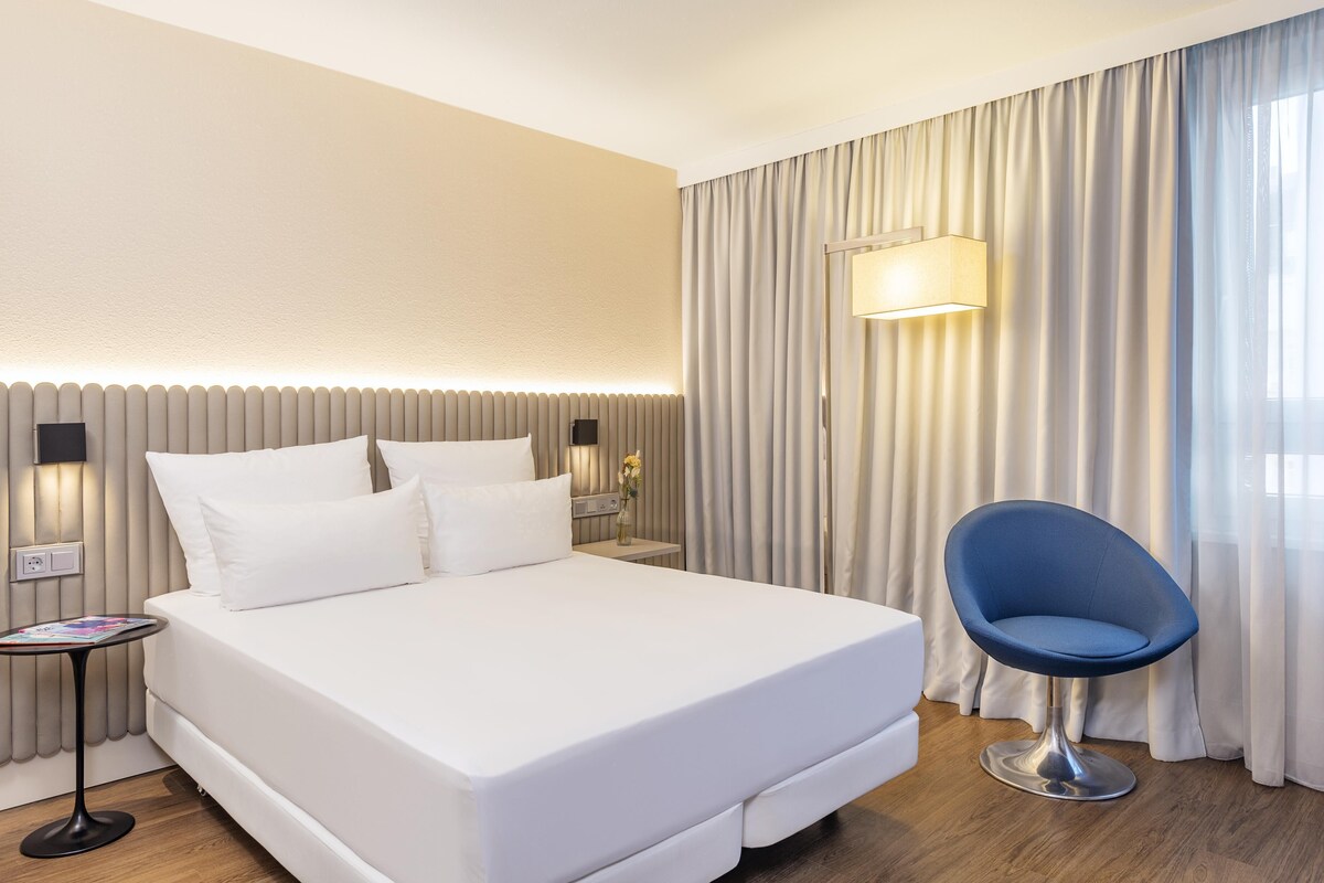 A hotel room with a bed, bedside table, lamp, chair, and curtains.