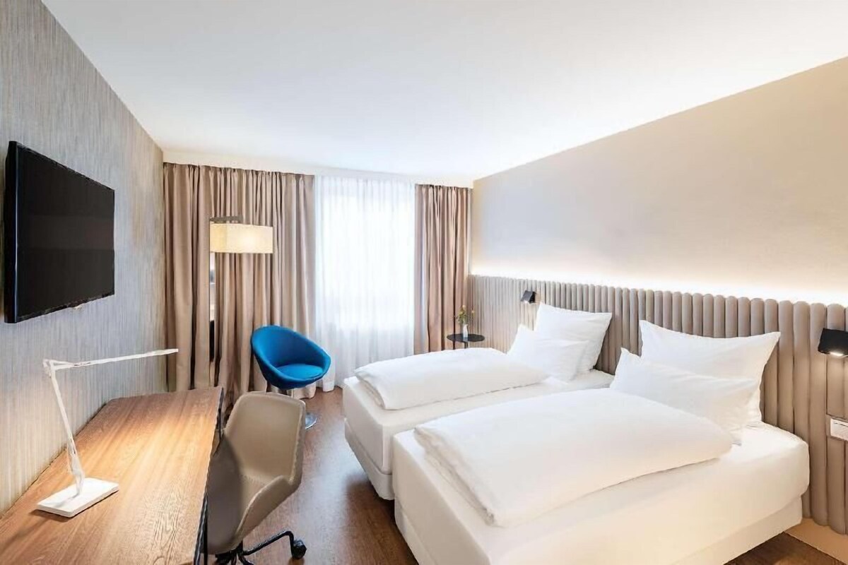 A hotel room with two beds, a desk with a chair, a TV mounted on the wall, and a window with curtains.