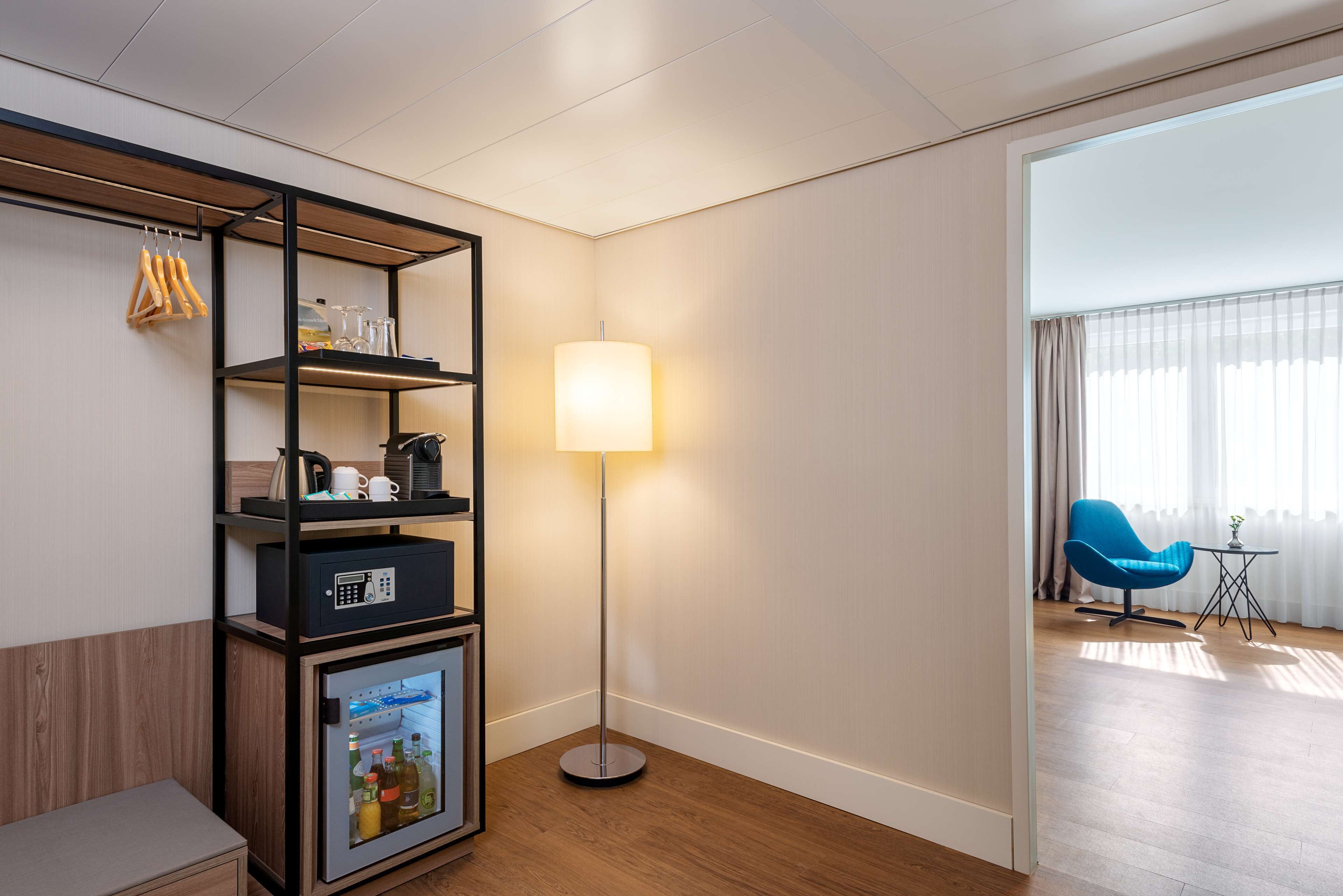 Ein Hotelzimmer mit Holzboden, einer Stehlampe, einem blauen Sessel und einem Minikühlschrank mit Getränken.