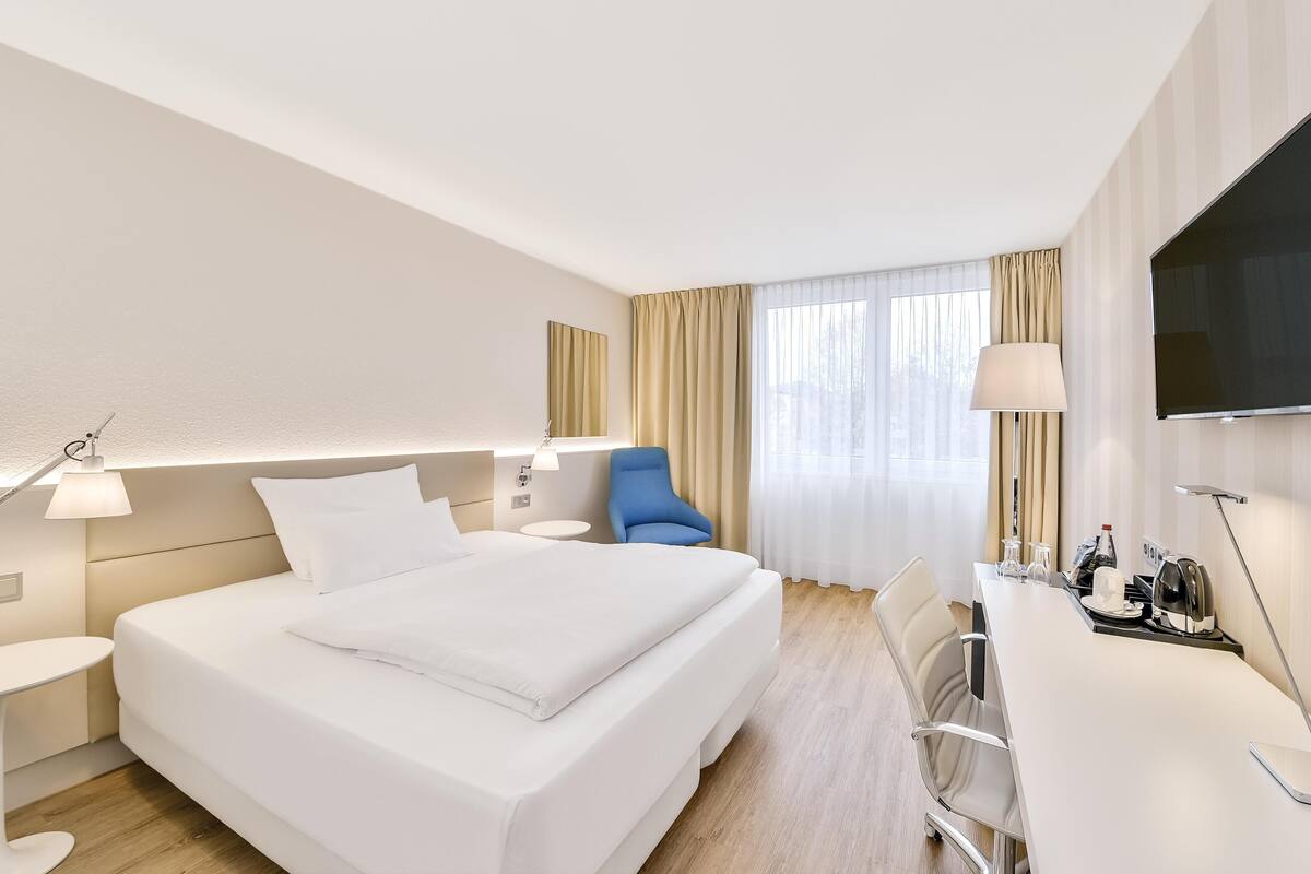 A hotel room with a large bed, a desk, a chair, a TV, and a window with curtains.