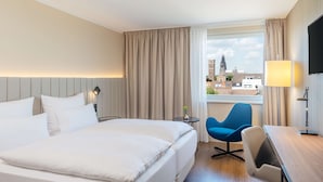 Ett hotellrum med en säng, ett skrivbord, en stol, en lampa och utsikt över en stad.