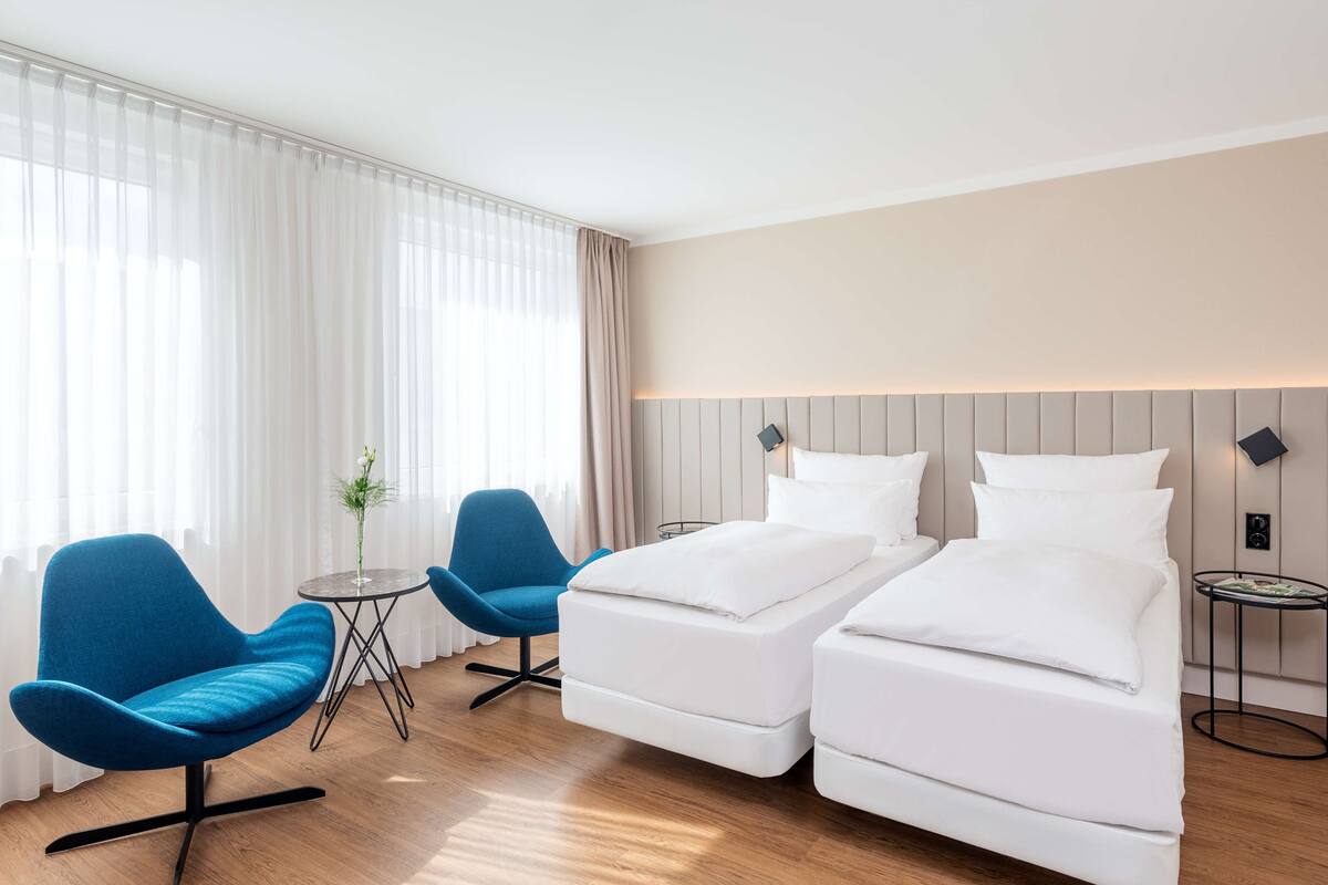 A hotel room with two single beds, a blue chair, a small round table, and a large window with sheer curtains.