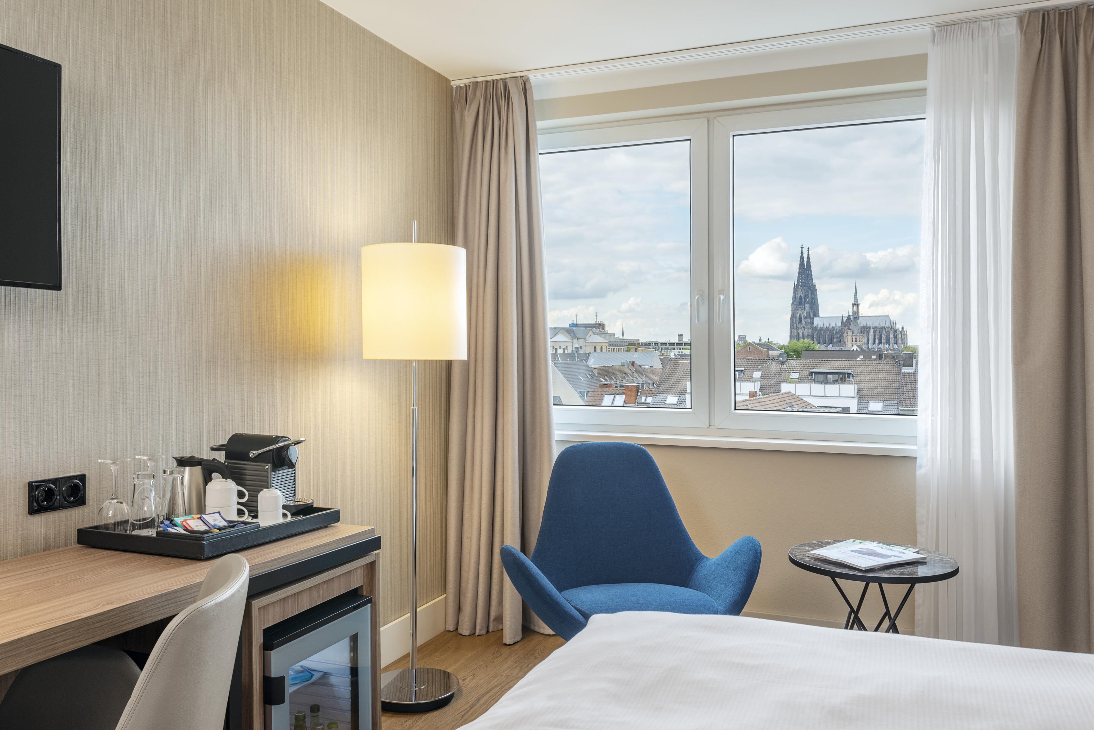 Habitación de hotel con cama, escritorio con cafetera, silla, mesita y vista a un paisaje urbano.
