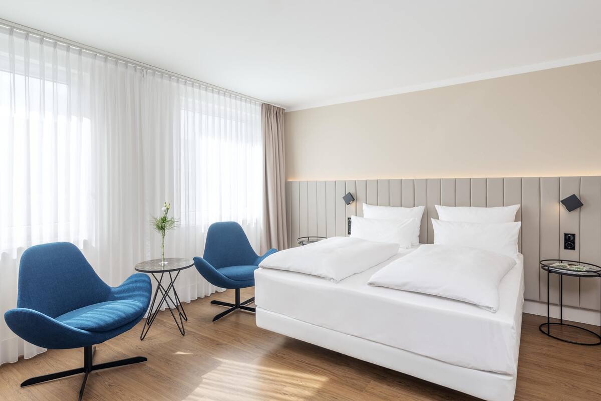 A hotel room with a large bed, two blue chairs, a small round table, and a potted plant.