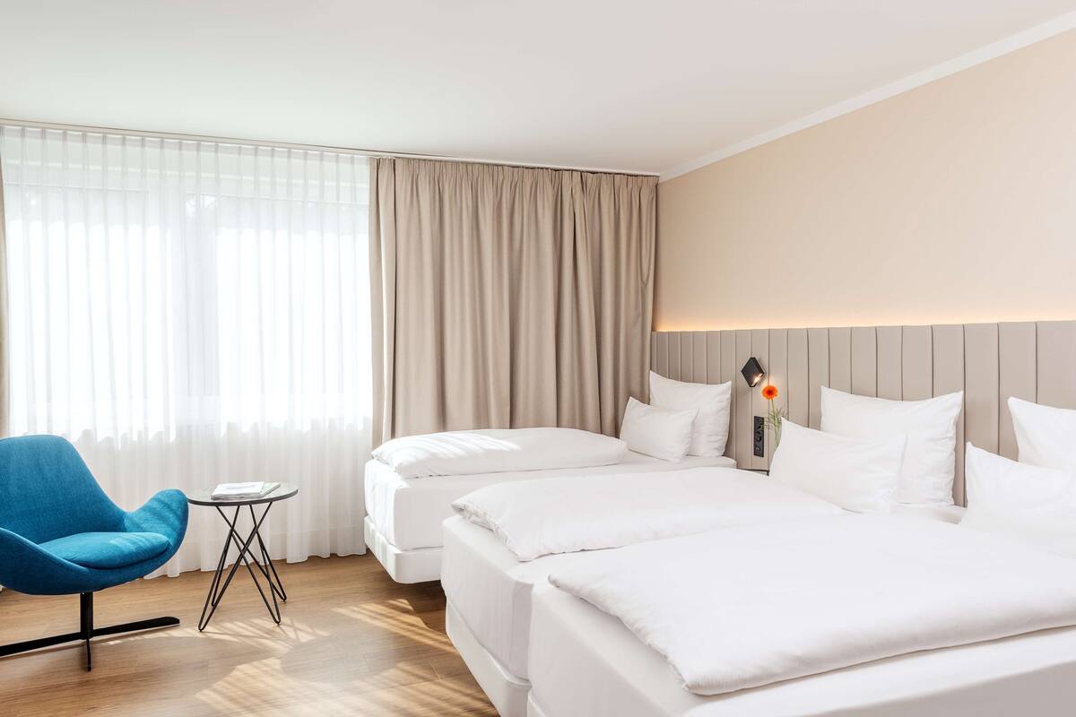 A hotel room with two beds, a blue armchair, a small table, and a nightstand with a lamp.