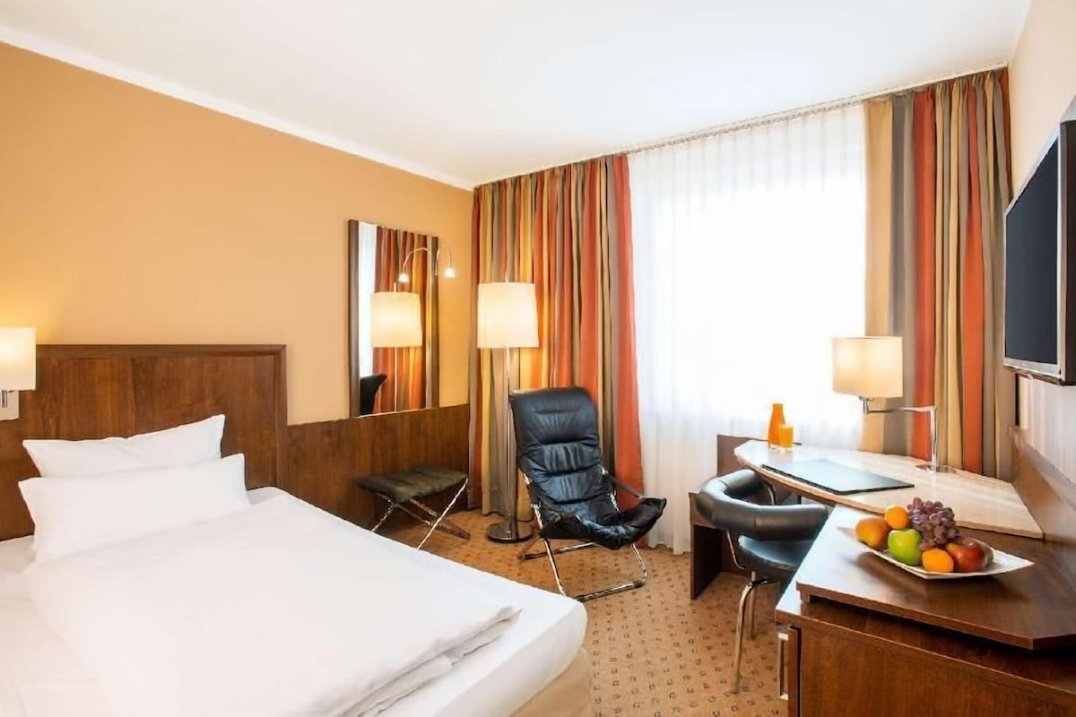 A hotel room with a bed, a desk with a chair, a television, and a window with curtains.