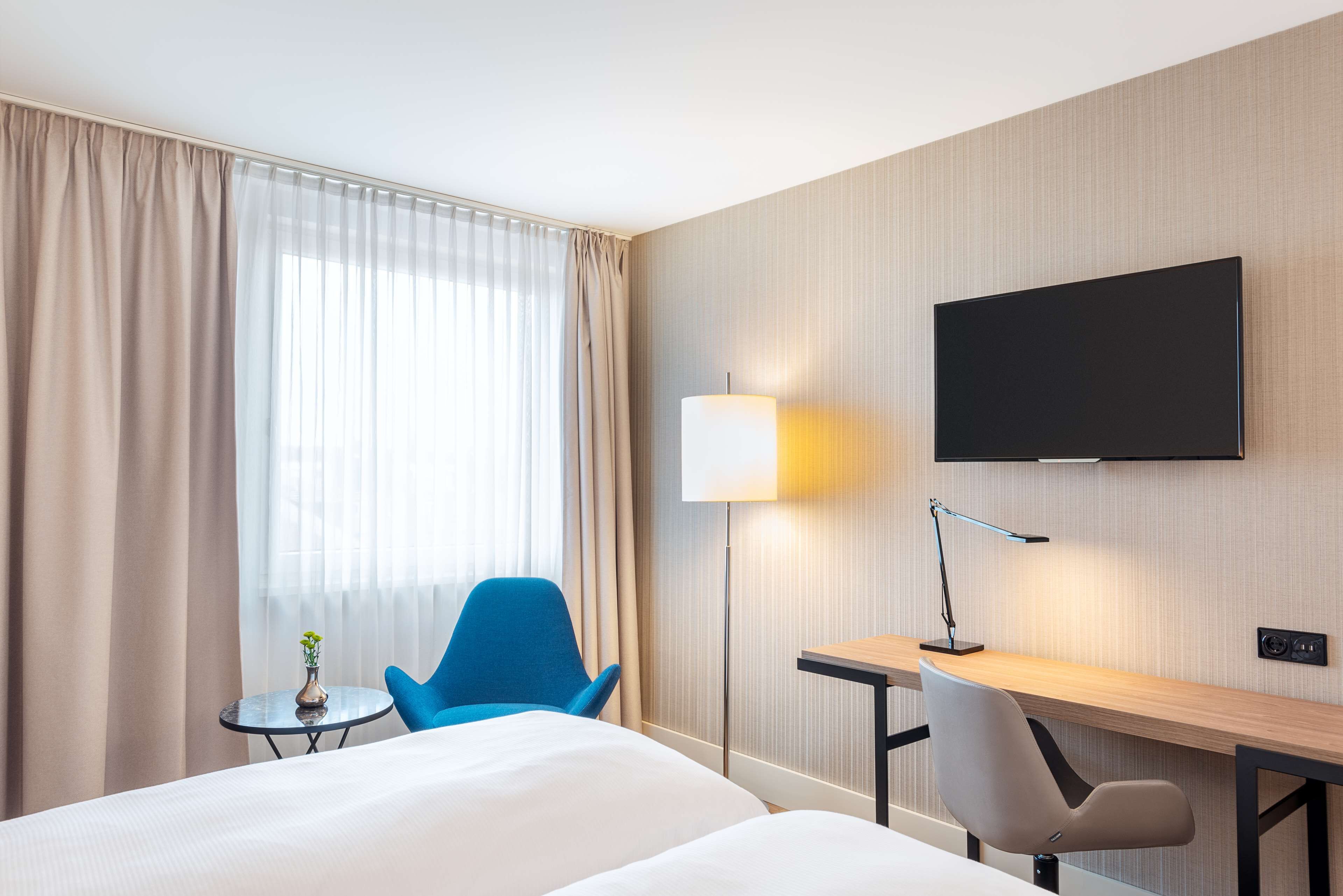 A hotel room with a bed, a desk with a lamp, a TV, and a chair.