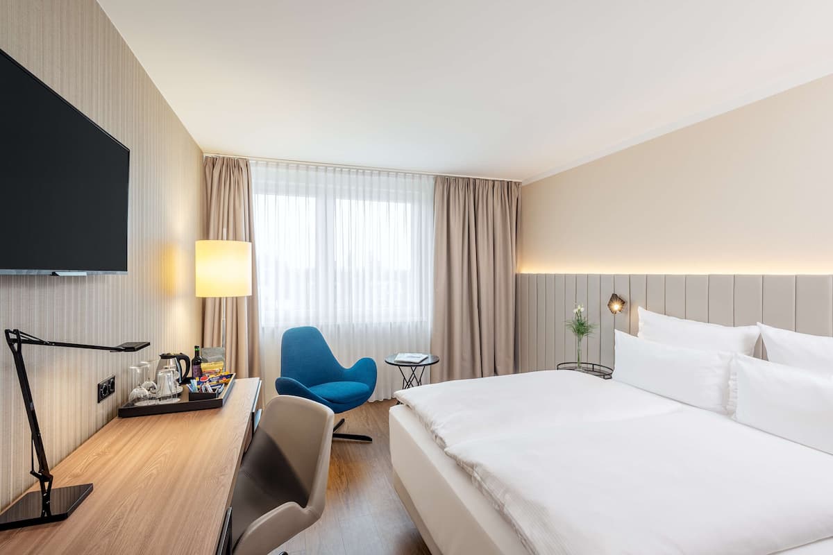 A hotel room with a large bed, a desk with a lamp, a TV, a blue chair, a small table, and a nightstand with a lamp.