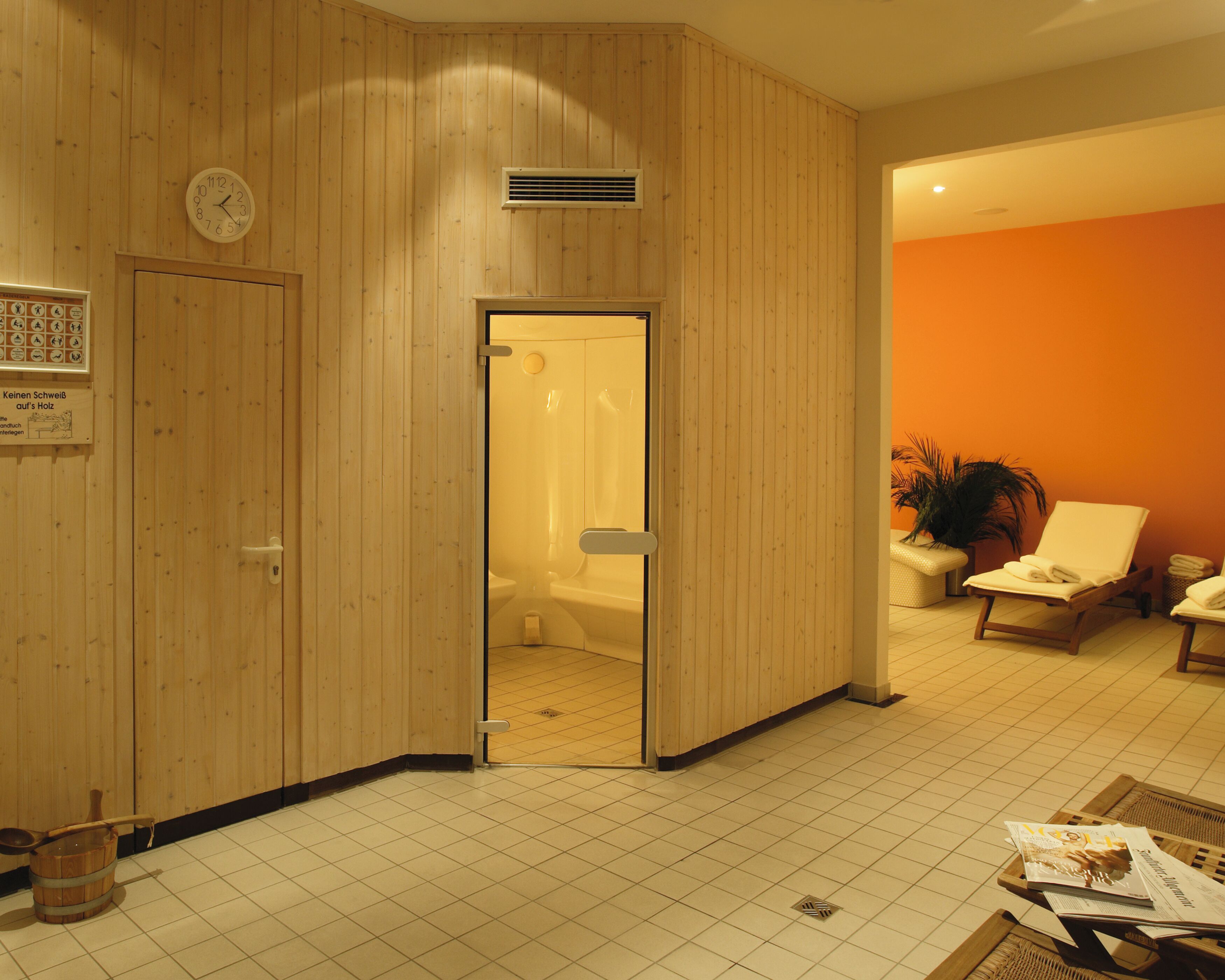 A room with a wooden panel wall, a clock, a shower area, a seating area with a chair and a plant, and a tiled floor.