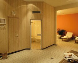 Ein Zimmer mit einer Holzwand, einer Uhr, einem Duschbereich, einer Sitzecke mit Stuhl und Pflanze sowie einem gefliesten Boden.