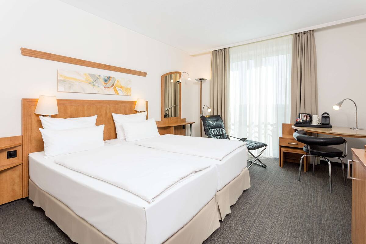A hotel room with a double bed, a desk with a chair, a lamp, and a large window with curtains.
