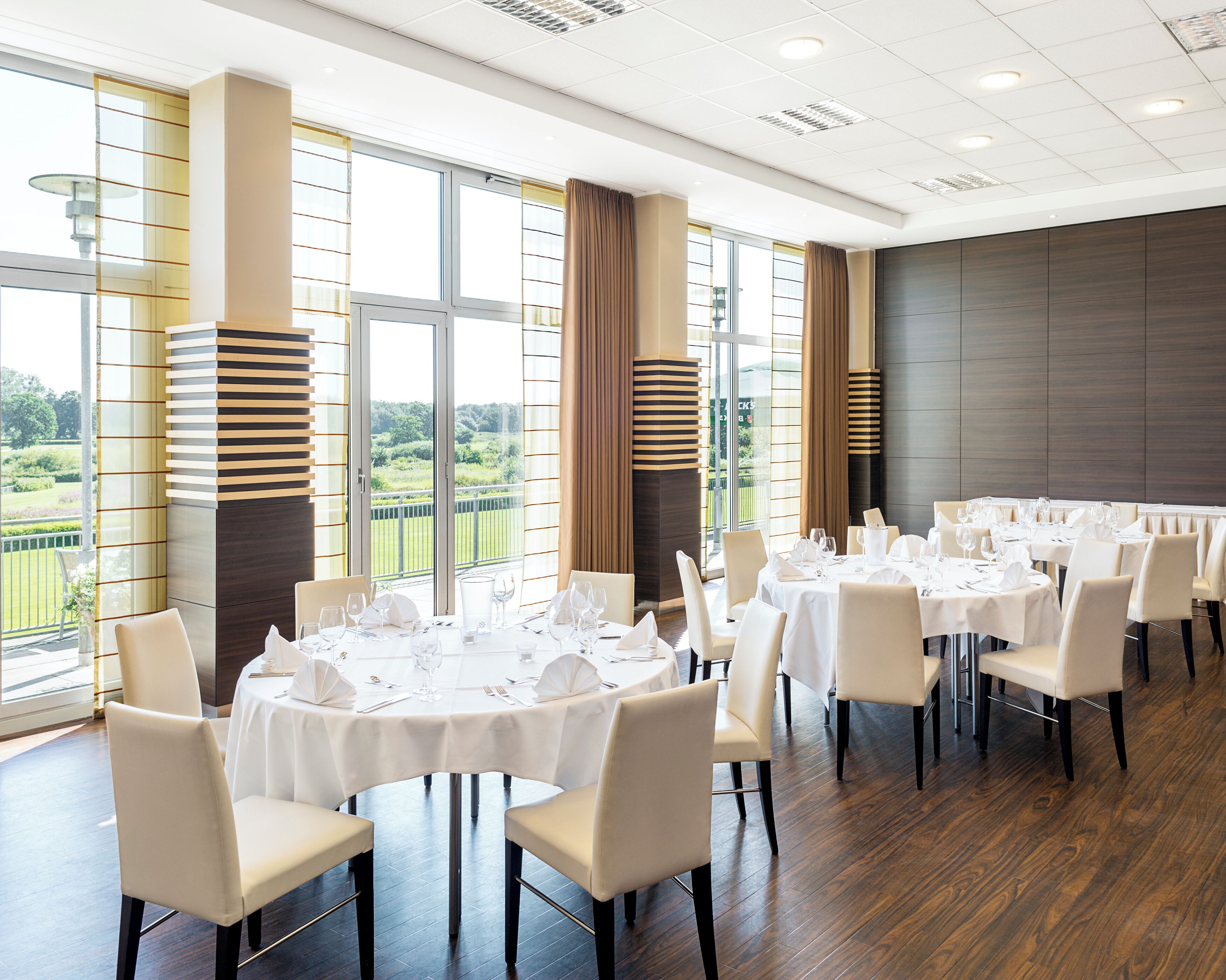 Un espace repas avec des tables rondes, des nappes blanches et des chaises, de grandes fenêtres donnant sur un paysage verdoyant et des luminaires modernes.